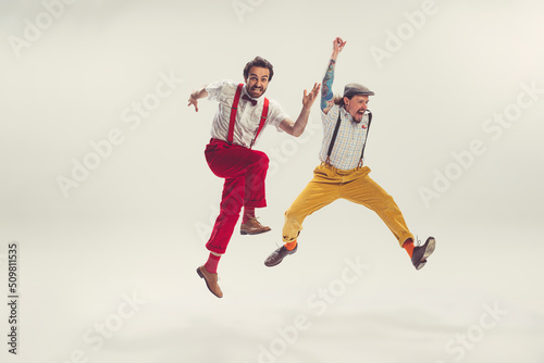 Two cheerfull dudes, young men in old-school fashioned attire running, jumping, having fun isolated on white background. Vintage, retro style concept