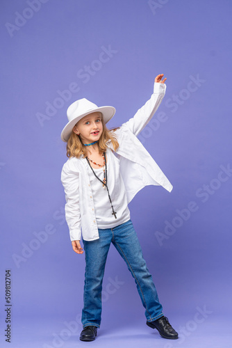 Portrait of happy cute little girl in a hat and unisex clothes on a purple background in full growth. stylish children's model is having fun and feels stunned, amazed. Advertising of children's goods © skif