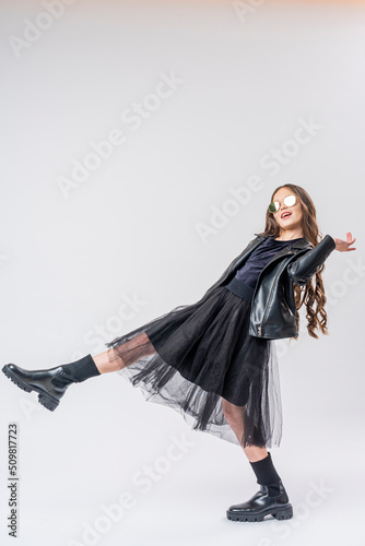 stylish teenage girl with dark hair, dressed in fashionable clothes, a leather jacket and sunglasses on white wall. full growth. concept of youth and clothing.