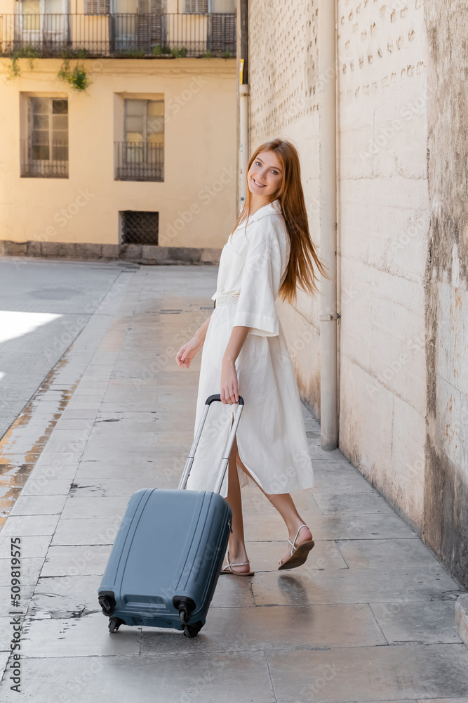 young joyful woman in dress walking with suitcase on street in valencia.