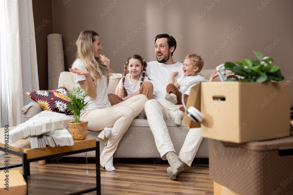 Loving family spends time with each other during move relaxing on couch, talking, discussing. Parents with two children enjoy their new apartment, cardboard boxes with unpacked belongings all around. Loving family spends time with each other during move relaxing on couch, talking, discussing. Parents with two children enjoy their new apartment, cardboard boxes with unpacked belongings all around.