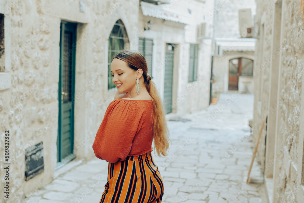 Beautiful stylish girl with long earrings, wearing an orange suit looks at the camera. Traveling around Europe Fashionable woman smiling in the old town and posing