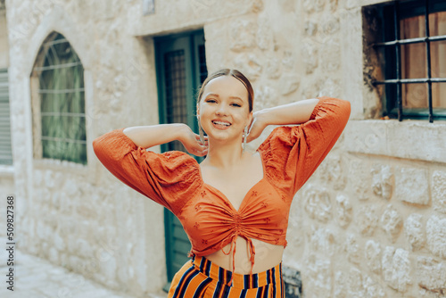 Fashionable woman smiling in the old town and posing. Beautiful stylish girl with long earrings, wearing an orange suit looks at the camera. Traveling around Europe. © MoreThanProd