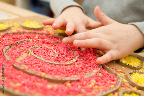 Boy plays with colored rice. Implement for children to develop fine motoric skills through play. DIY toy to learn hand - eye coordination. Montessori type pre school activities at home.