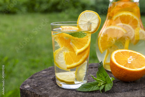 Citrus drink with lemon, orange and mint. Healthy and healthy drink in a transparent glass on a stump against the background of green grass with copy space