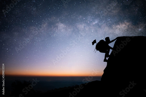 A young tourist with backpack is climbing a cliff alone with beautiful view of night sky, star and Milky Way. He had an effort to climb all the way to the top of the mountain and he had to succeed.