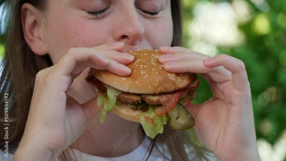 Beautiful young woman eating tasty burger outdoors, closeup. Slow motion effect Beautiful young woman eating tasty burger outdoors, closeup. Slow motion effect