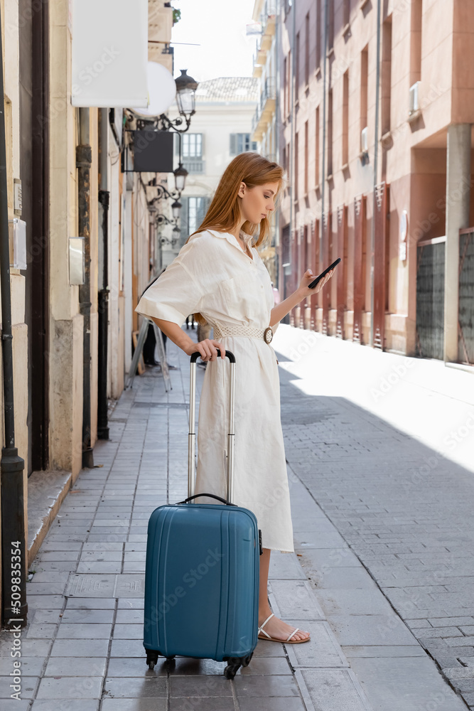 redhead woman in dress using smartphone and standing with luggage on urban street in valencia.