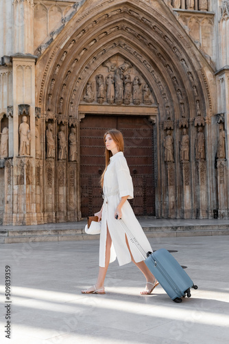 full length of redhead young woman in dress holding purse and walking with luggage in valencia.