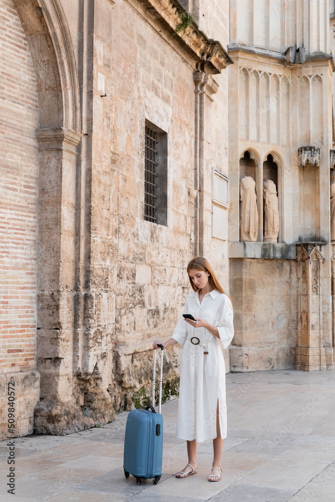 full length of redhead woman standing with luggage and using smartphone while searching geolocation in valencia.