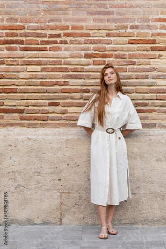 full length of dreamy redhead woman in dress posing near brick wall on european street.