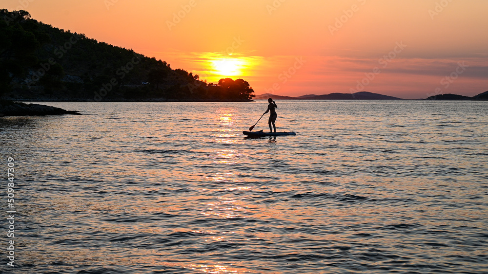 Fototapeta premium Silhouette of woman surfing on stand up paddle board on the sea. Woman enjoying on Paddle-board at summer. Paddling on the quiet sea at sunset. SUP