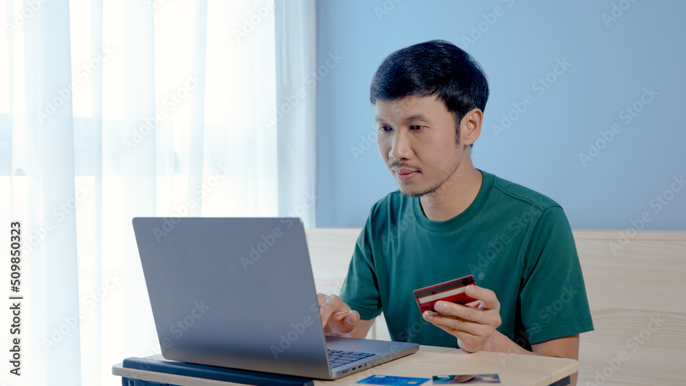 Handsome Asian man, wearing a green shirt, looks happy when he successfully pays for online purchase via a laptop computer with a credit card, online shopping. Handsome Asian man, wearing a green shirt, looks happy when he successfully pays for online purchase via a laptop computer with a credit card, online shopping.