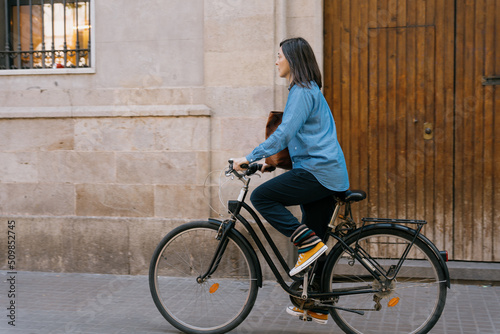 A woman with a blue shirt and yellow sneakers riding her black urban bicycle in the city of Barcelona. An old building with a wooden door and a stone wall in the background.