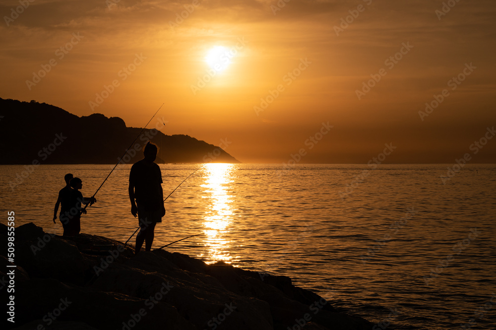 SILHOUETTE OF FISHERMEN AT SUNSET ON THE CLIFFS BY THE SEA WITH BOAT IN THE BACKGROUND