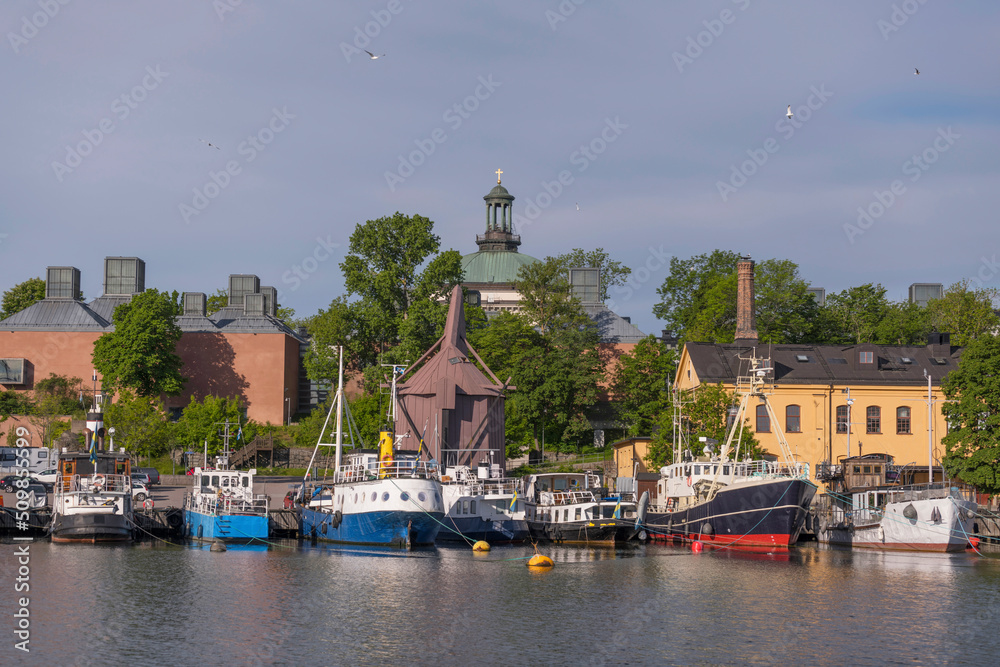 Fototapeta premium Small ships and boats at a pier on the island Skeppsholmen, a church tower and old military buildings sunny summer morning in Stockholm