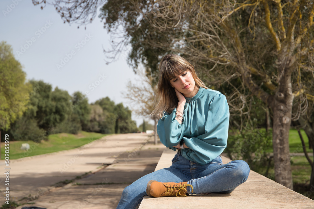 Young, pretty, blonde woman in green shirt, jeans and brown boots, sitting with serious gesture in a park. Concept depression, problems, seriousness, concern. Young, pretty, blonde woman in green shirt, jeans and brown boots, sitting with serious gesture in a park. Concept depression, problems, seriousness, concern.