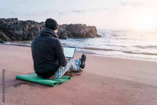 digital nomad working on his laptop outdoors from the beach at sunset, back view