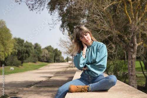 Young, pretty, blonde woman in green shirt, jeans and brown boots, sitting with serious gesture in a park. Concept depression, problems, seriousness, concern.