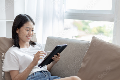 Asian woman sitting on the sofa in the living room using a tablet, Looking at tablet screen, Relaxing at home, Working through the Internet communication system, Happy working lifestyle. Free Download Asian woman sitting on the sofa in the living room using a tablet, Looking at tablet screen, Relaxing at home, Working through the Internet communication system, Happy working lifestyle. - GFXLoad