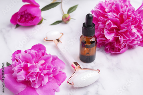 Glass bottle with oil, roller for face massage on marble background with peony flowers