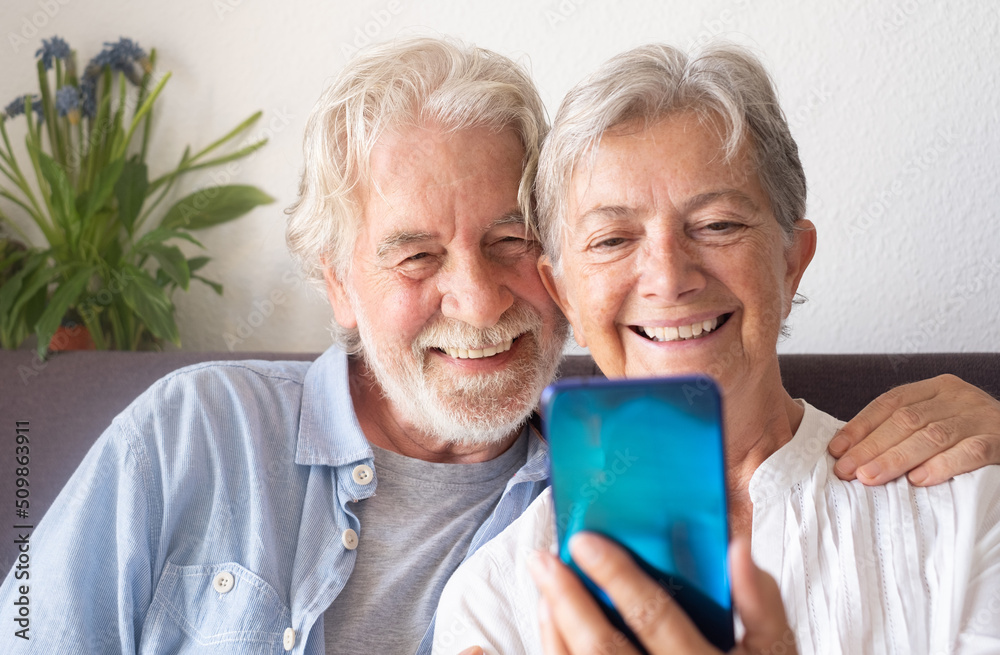 Portrait of beautiful happy caucasian senior couple of grandparents, looking at mobile phone smiling, hugging and resting on sofa at home Portrait of beautiful happy caucasian senior couple of grandparents, looking at mobile phone smiling, hugging and resting on sofa at home