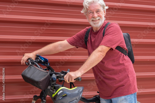 Smiling senior cyclist man in urban city with his electric bicycle close to a red metal panel. Healthy lifestyle for retired people and sustainable mobility concept