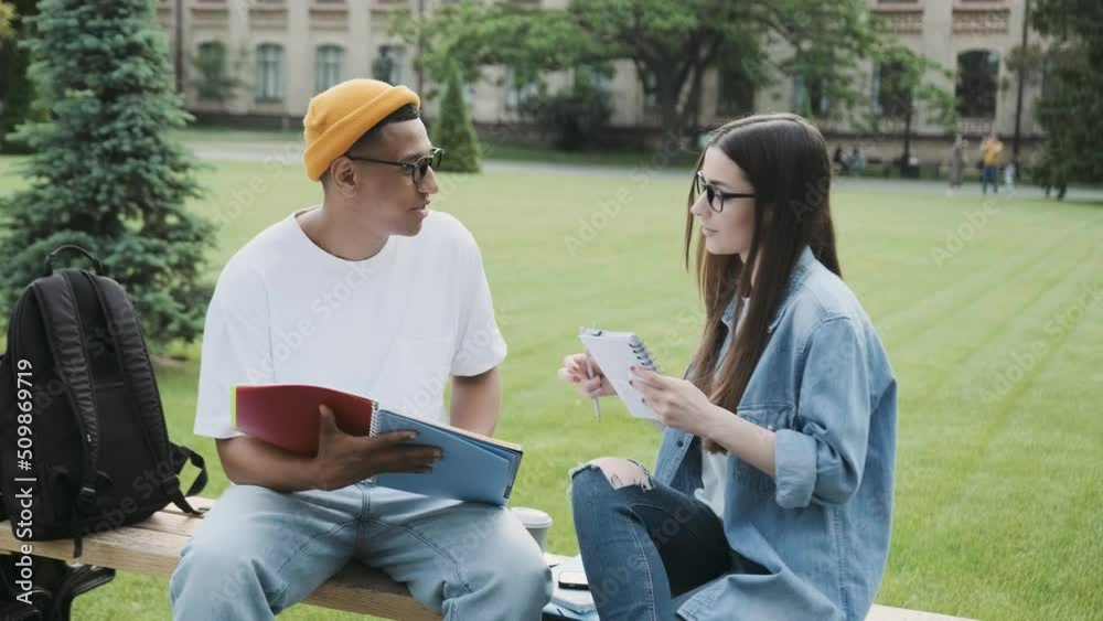 Couple of international students working on a university project in the campus, sitting on a bench.