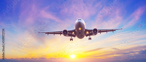 Panoramic view of the silhouette of an aircraft landing with extended landing gear, sky with different types of clouds gradient illuminated by the evening sun leaving the horizon.