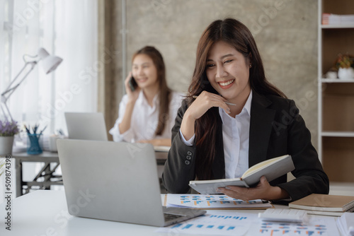 Young business woman working on laptop and writing on note book with happy and smile face with co-worker is talking via mobile phone behind her in office spec.