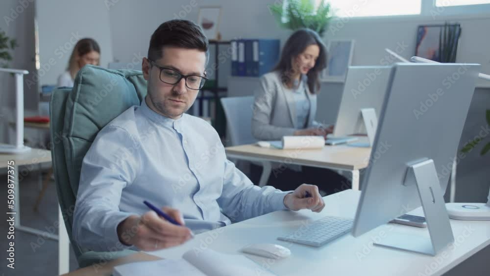 Businessman with glasses working on computers writing on notes in the office. In the background businesspeople work. Slow-motion Businessman with glasses working on computers writing on notes in the office. In the background businesspeople work. Slow-motion
