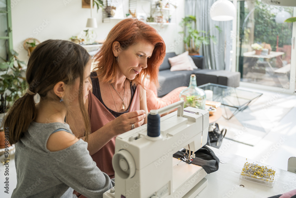 Female designer sewing new clothes while sitting at her workplace at home with daughter Female designer sewing new clothes while sitting at her workplace at home with daughter