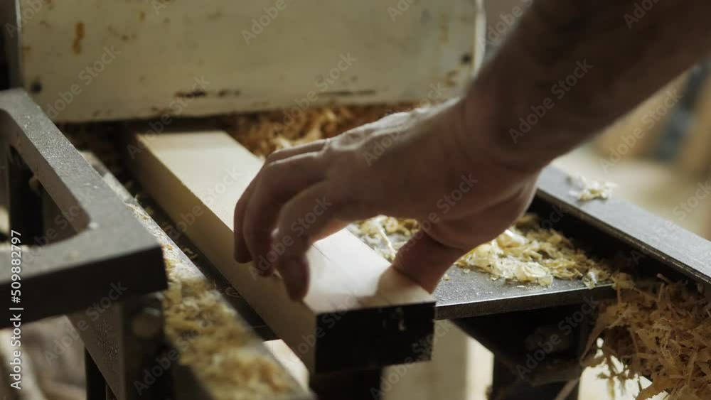 A carpenter in the workshop works on a woodworking machine, processes wood for the manufacture of furniture parts. The board is shoved into the machine for processing in natural light. Close-up