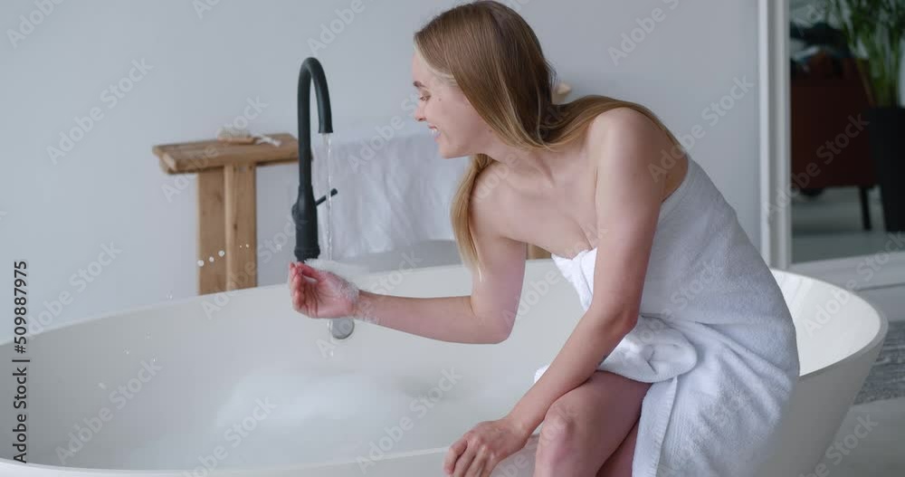 Pretty woman with long blond hair sits on the edge of a bathtub, wrapped in white terry towel, trying the water for temperature, taking full bath with soap foam. Freshness, purity, body care concept