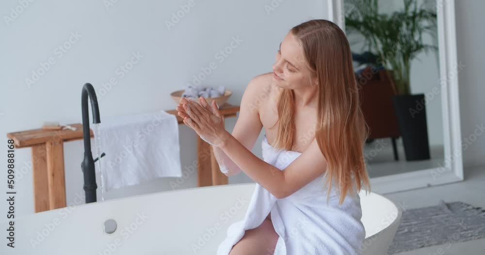 Adorable blonde woman sitting on the edge of a bathtub, wrapped in white terry towel, trying the water for temperature, preparing full bath with soap foam to relax at her day off. Body, skin care. Adorable blonde woman sitting on the edge of a bathtub, wrapped in white terry towel, trying the water for temperature, preparing full bath with soap foam to relax at her day off. Body, skin care.