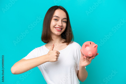 Young Ukrainian woman holding a piggybank isolated on blue background with thumbs up because something good has happened