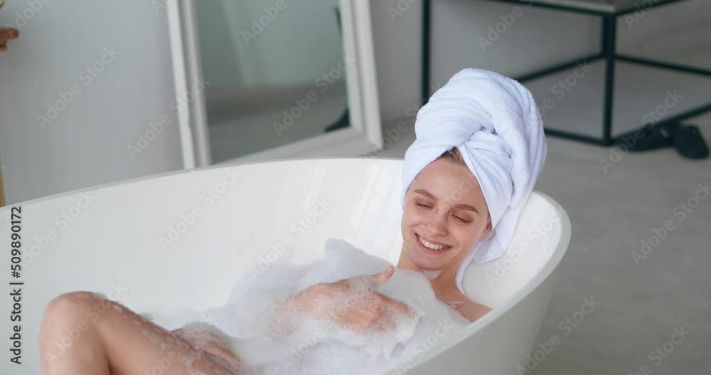 Beautiful woman with a bath towel on head lying on a bathtub, smiling, relaxing, massaging her body while taking a bubble bath with foam at white home bathroom, enjoying day off. Body care concept Beautiful woman with a bath towel on head lying on a bathtub, smiling, relaxing, massaging her body while taking a bubble bath with foam at white home bathroom, enjoying day off. Body care concept