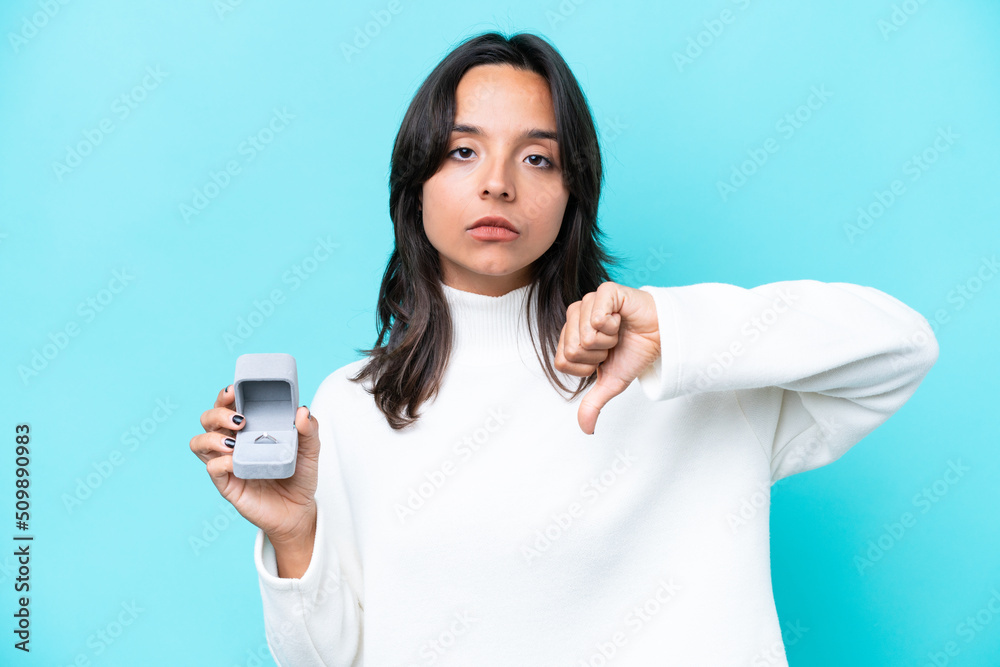 Young hispanic woman holding a engagement ring isolated on blue background showing thumb down with negative expression Young hispanic woman holding a engagement ring isolated on blue background showing thumb down with negative expression