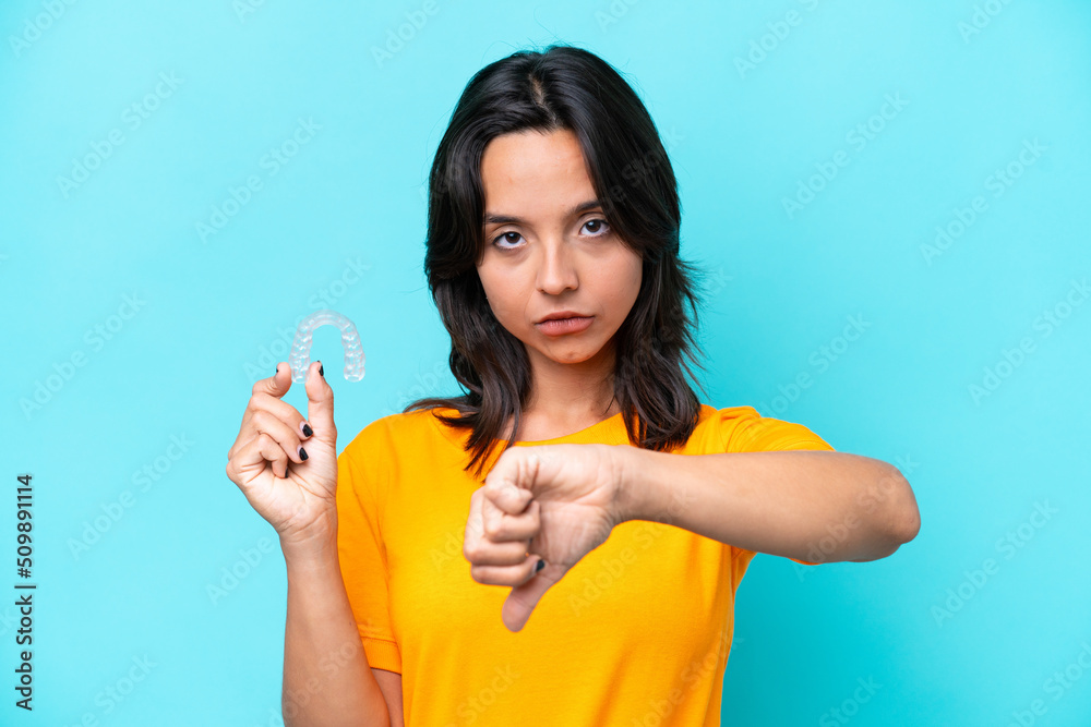 Young hispanic woman holding invisible braces isolated on blue background showing thumb down with negative expression Young hispanic woman holding invisible braces isolated on blue background showing thumb down with negative expression
