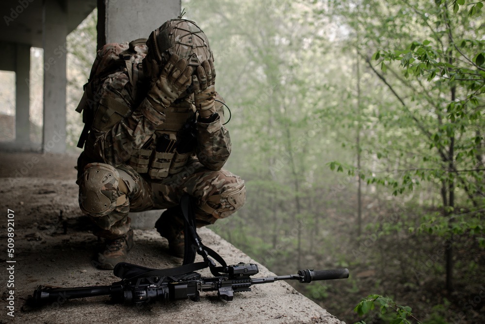 Military soldier in uniform sitting on his knees, releasing his head in front of the machine gun. Loss of a combat partner. Defeat in battle Military soldier in uniform sitting on his knees, releasing his head in front of the machine gun. Loss of a combat partner. Defeat in battle
