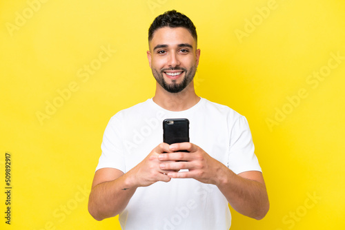 Young Arab handsome man isolated on yellow background looking at the camera and smiling while using the mobile © luismolinero