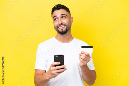 Young Arab handsome man isolated on yellow background buying with the mobile with a credit card while thinking