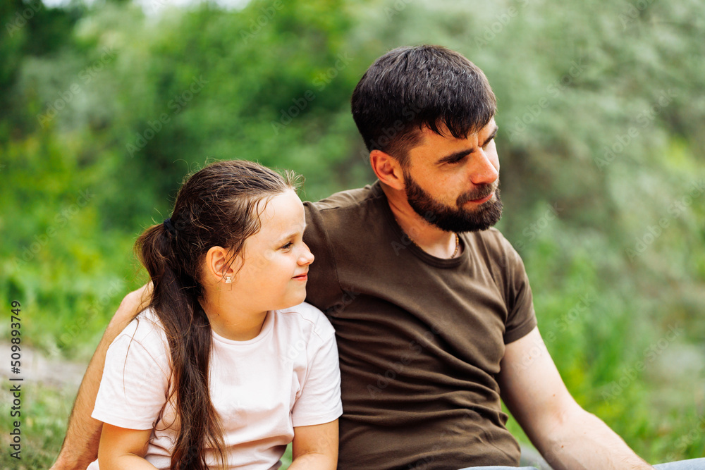 Portrait of satisfied family sitting on picnic in park forest around trees bushes. Happy middle-aged man father embracing little girl daughter, having fun. Love, family, summer, togetherness, care. Portrait of satisfied family sitting on picnic in park forest around trees bushes. Happy middle-aged man father embracing little girl daughter, having fun. Love, family, summer, togetherness, care.