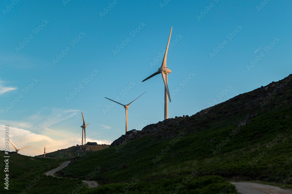 Turbinas eólicas na paisagem montanhosa ao pôr do sol com uma estrada sobre a montanha - obrazy, fototapety, plakaty 