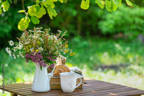 Bouquet of meadow flowers, croissant, cup of tea or coffee, books on table in summer garden. Rest in garden, reading books, breakfast, vacations in nature concept. Summertime in garden on backyard photo