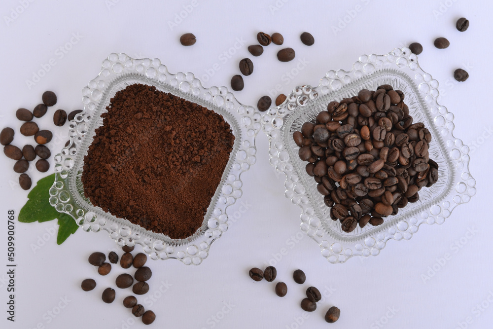 Aerial photo of two small glass plates filled with coffee beans and ground coffee decorated with green leaves Aerial photo of two small glass plates filled with coffee beans and ground coffee decorated with green leaves