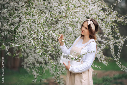 A beautiful young woman in a retro apron enjoys life in a spring blooming garden with a background of flowering trees. A girl among the apple blossoms. Spring in the countryside concept.