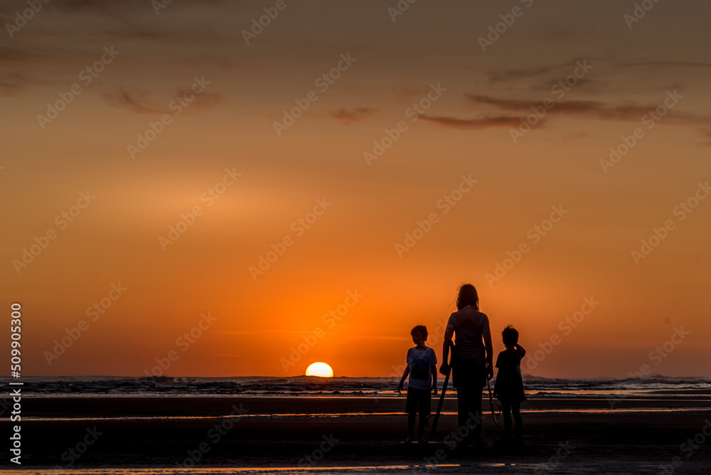 Backlight of woman on crutches with two children watching sunset over the sea from the beach