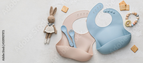Silicone baby bibs with plastic cutlery and toys on light background with space for text