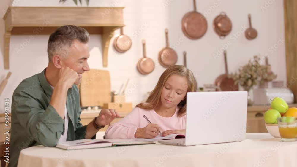 Happy caucasian father using laptop and helping his preteen beautiful daughter with homework at home. Cute child writing in notebook with confident dad at kitchen table. Happy caucasian father using laptop and helping his preteen beautiful daughter with homework at home. Cute child writing in notebook with confident dad at kitchen table.